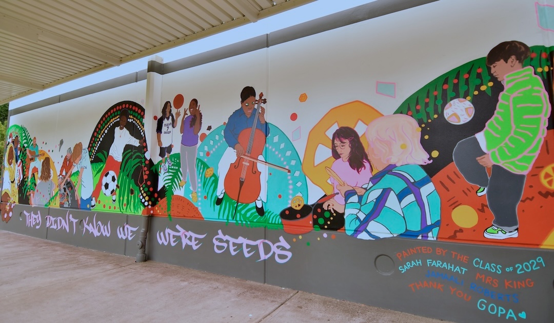 Colorful mural on a school wall featuring students engaged in creative activities such as playing the cello, painting, reading, and playing soccer. The background is filled with vibrant patterns, greenery, and geometric shapes. Text on the mural reads: They didnt know we were seeds. Painted by the class of 2029 Sarah Farahat Mrs King Jamaali Roberts Thank you GOPA. The scene feels joyful, collaborative, and energetic, celebrating diversity and learning in a lively school environment.